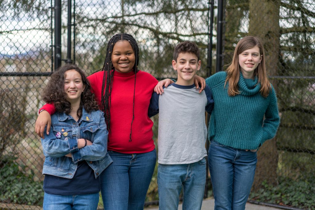 Four teenagers stand side by side outdoors, smiling at the camera. They are dressed casually in jeans and sweaters, with their arms around each other in front of a chain-link fence and trees.