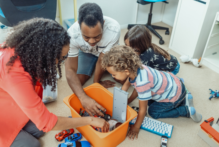 Two adults and two children kneel on the floor, organizing or playing with toys from an orange storage bin in a brightly lit room. A toy keyboard and several toy cars are visible nearby.