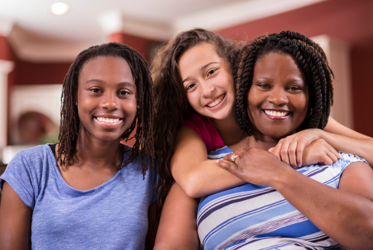 Three people are smiling indoors. Two are seated, and one stands behind them with her arms around their shoulders. All look happy and are close together, suggesting a warm, family or friendship bond.