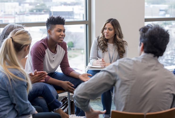 A diverse group of people sits in a circle indoors, engaged in a discussion. One person speaks while others listen attentively. Large windows show an urban view in the background.